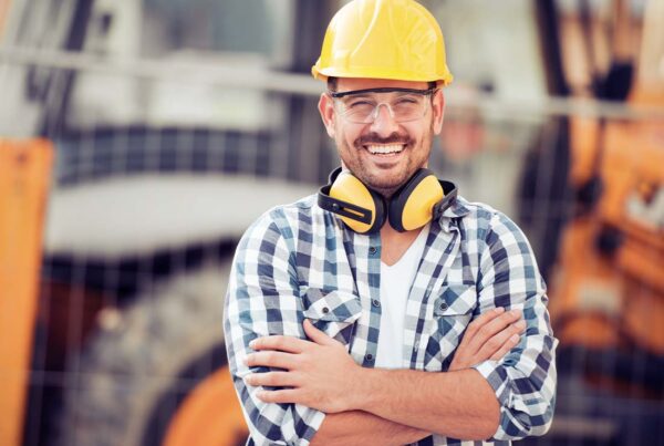 Contractor Insurance - Portrait of a Smiling Young Construction Worker in a Hard Hat with Arms Crossed Standing in Front Construction Equipment at a Construction Site