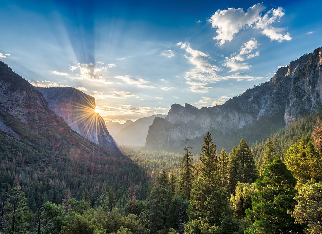 About Our Agency - Scenic View of the Sun Setting Over the Mountains and Surrounding Green Trees with a Cloudy Blue Sky in a State Park