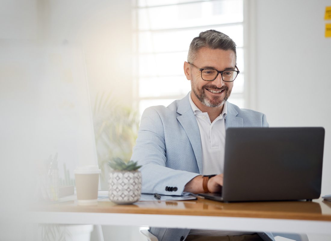 Blog - Older Man Smiling While Looking at Their Laptop and Typing in Their Office on a Nice Day